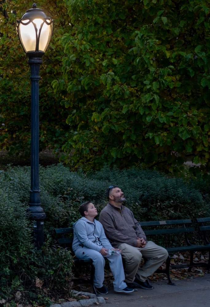 Two people on a park bench under a lit lamppost at dusk