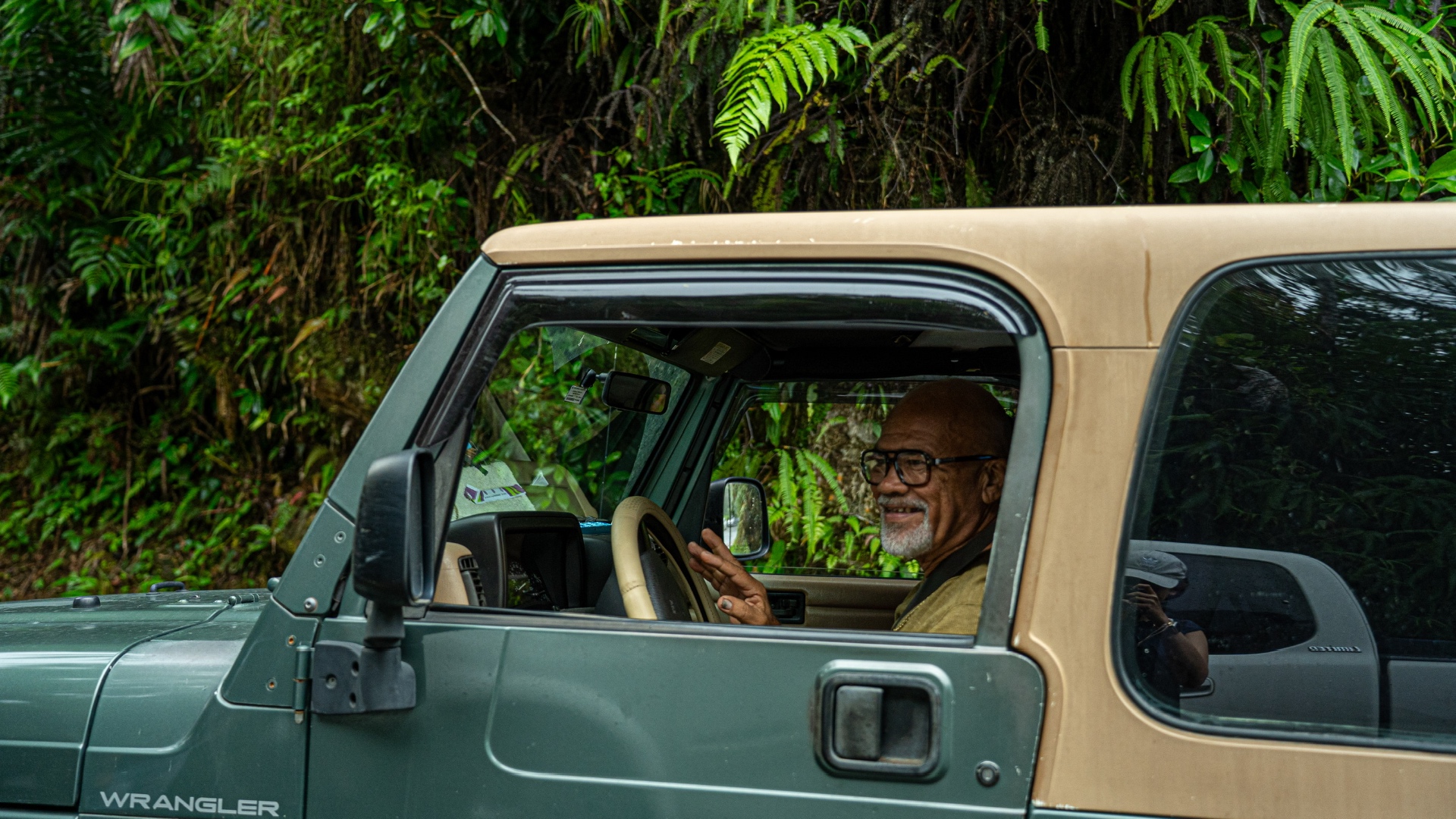 A smiling man waving from the window of his Jeep in the rainforest